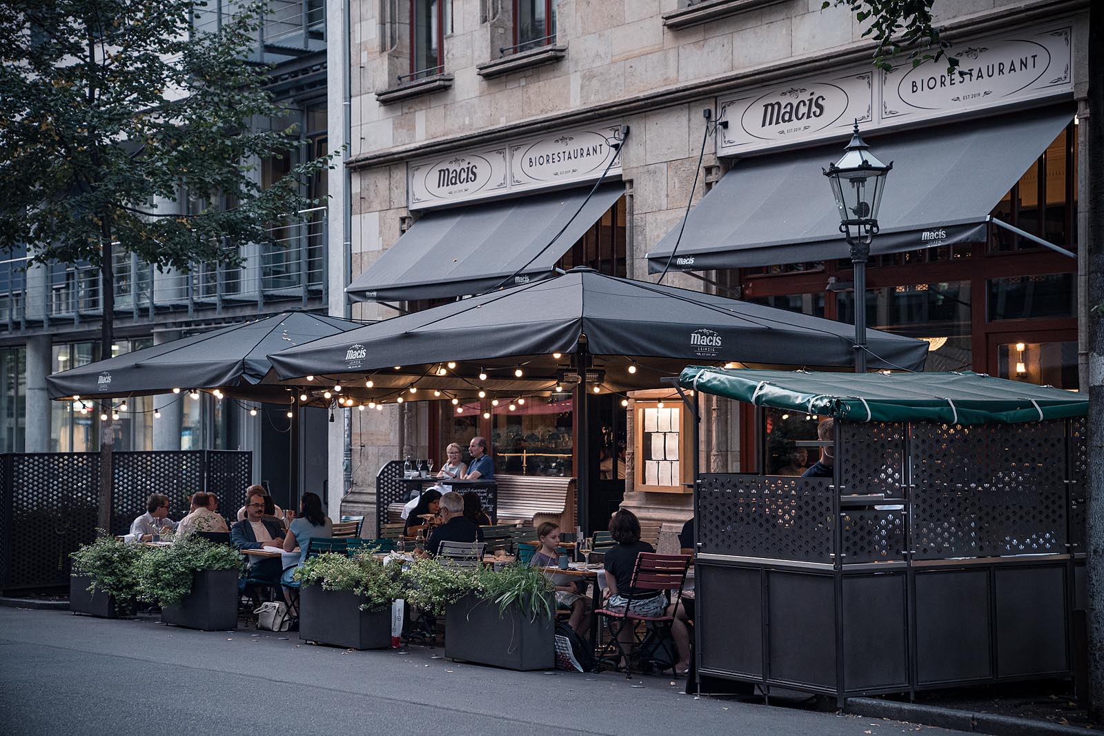 Abendaufnahme des Restaurants von außen, Gäste sitzen unter Lichterketten auf der Terrasse des macis Leipzig.